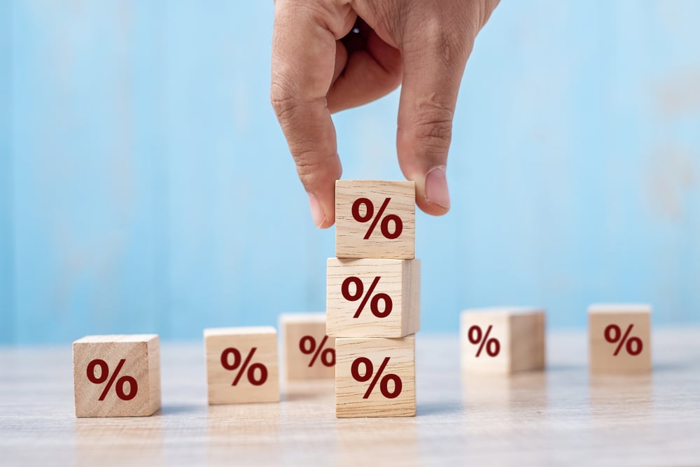A hand placing a wooden block with a percentage symbol on top of a stack of similar blocks, representing rising interest rates or financial growth.