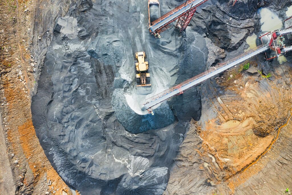 An aerial view of a mining site featuring a yellow front loader moving dark grey material near conveyor belts and large piles of extracted earth.